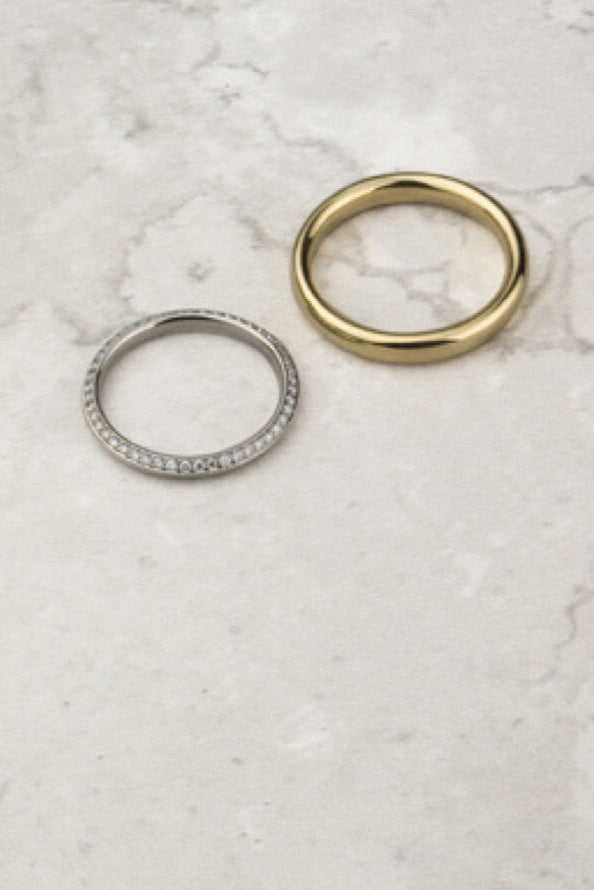A still life picture of two rings on a marble background. One heavy wedding band is made in yellow gold. The other is an eternity ring made in platinum with diamonds.